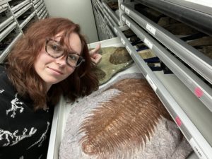 A young woman with reddish hair next to a museum specimen drawer with a trilobite