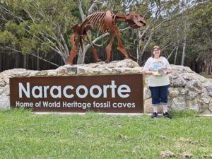 A statue of Diprotodon on a rock behind a sign that says "Naracoorte'. A woman stands next to the sign holding up a copy of a children's book.
