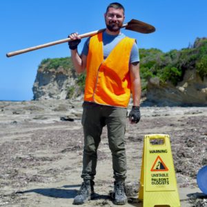 A white, male, youngish palaeontologist wearing a high visibility jacket and holding a shovel next to a sign that says 'Caution: Unstable. Palaeontologists'.